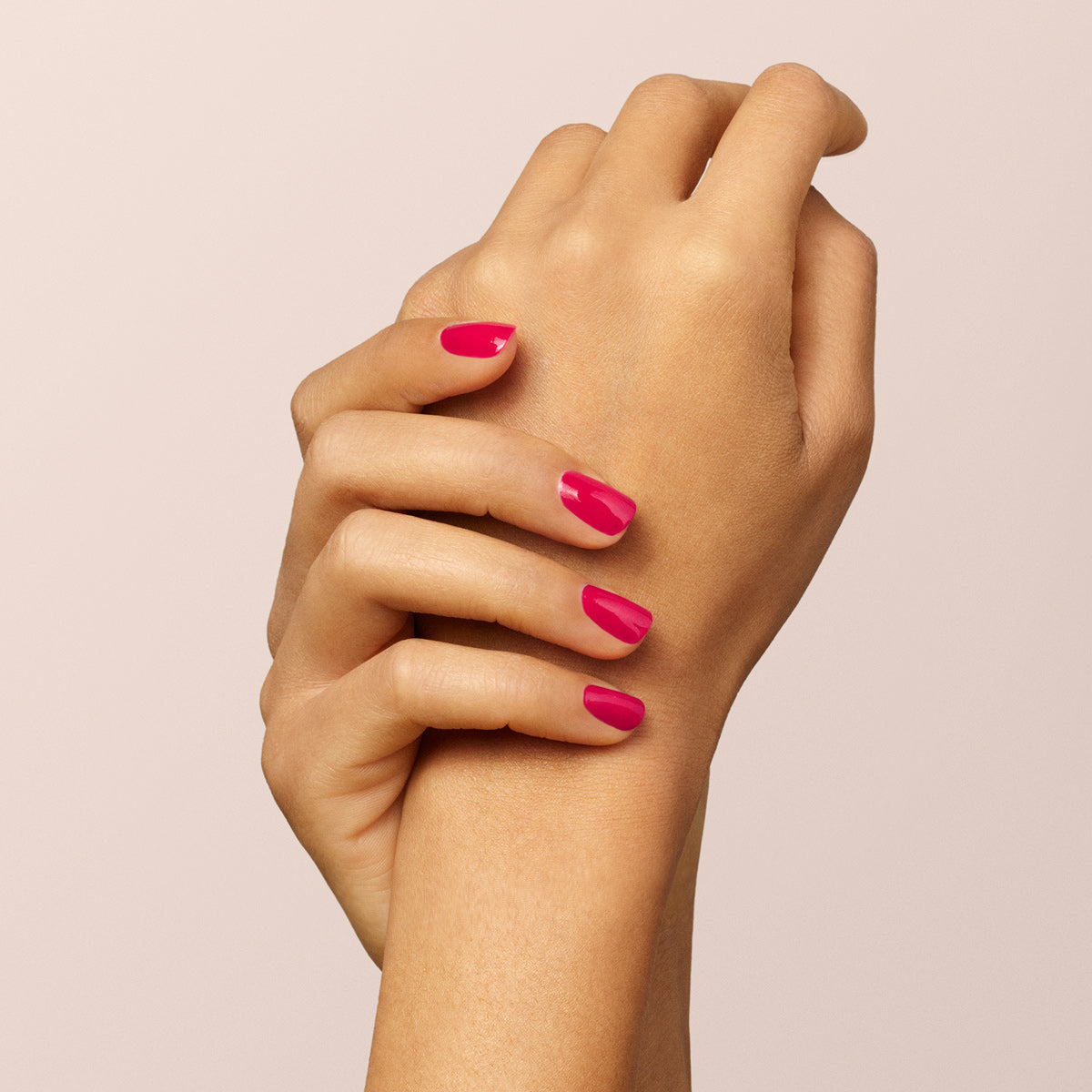 A close-up of two hands with neatly manicured nails painted in Péonie by Manucurist, a vibrant raspberry pink shade, set against a soft beige background.