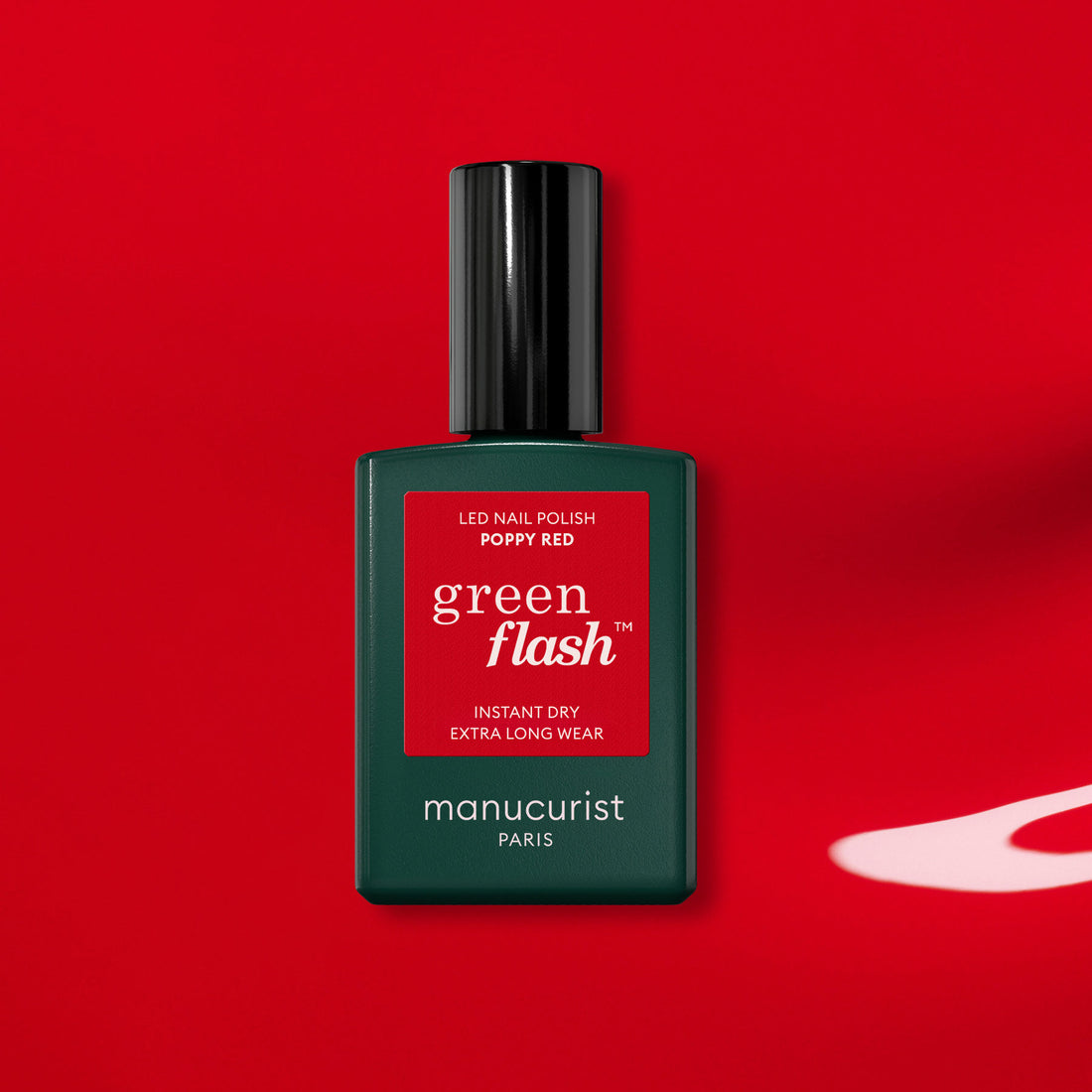 A bottle of Manucurist Poppy Red gel polish with a black cap rests on a bold red background, casting a dramatic shadow.
