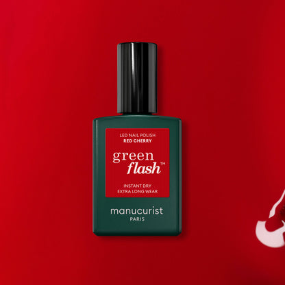 A bottle of Manucurist Red Cherry gel polish sits on a red background. The dark green bottle features a black cap and displays the product name on a red label.