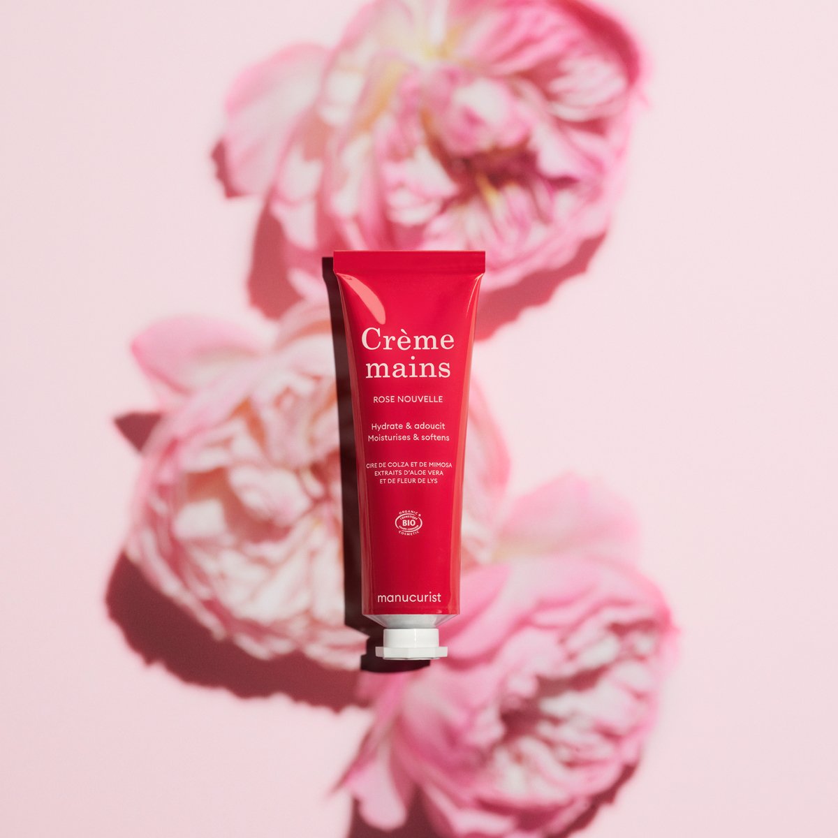 A red tube of Everyday Rose Hand Cream - Cosmos Organic by Manucurist sits centered on a pink background with three soft-focus pink flowers casting shadows. The product features a delightful rose scent.