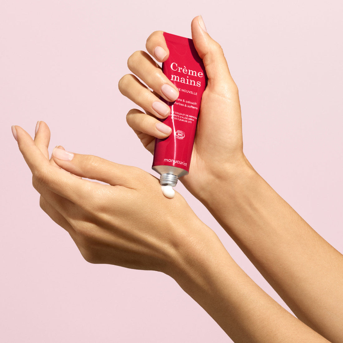 A person squeezes Manucurist Duo Rose Hand Cream  Rose Balm - Cosmos Organic from a red tube labeled Crème mains onto their hand, with a light pink background. This cream helps keep skin soft and hydrated.