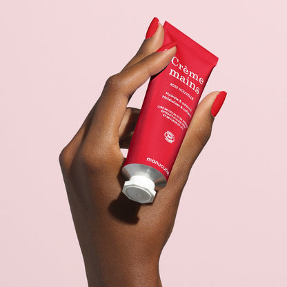 A hand with red-painted nails holds a red tube of Manucurist Everyday Rose Hand Cream - Cosmos Organic, featuring white text and a white cap, against a light pink background. The moisturizing cream has a subtle rose scent.