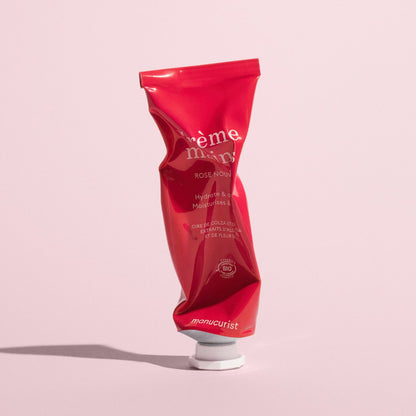 A red, crumpled tube of Manucurist Everyday Rose Hand Cream - Cosmos Organic with a white cap stands upright on a pink background. The tube displays “crème mains,” “Hydrate  Moisturise,” and other product details.