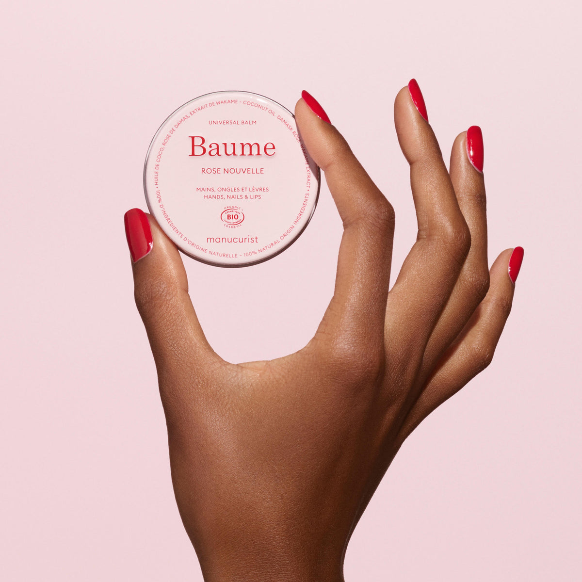 A hand with glossy red nails holds a round container of Manucurist Duo Rose Hand Cream  Rose Balm - Cosmos Organic—ideal for hands, nails, and lips—against a light pink background.