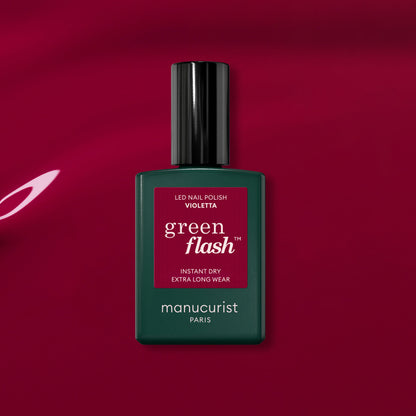 A bottle of Manucurist Violeta gel polish rests on a glossy magenta surface, light reflecting off the background to highlight its vibrant pink-purple tones.